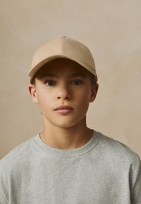 Berretto in tessuto beige con corona arrotondata e visiera curva, indossato da un bambino con una maglietta grigia a girocollo, su sfondo neutro.