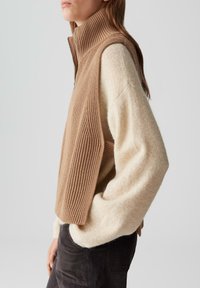 Brauner Strickwesten mit hohem Kragen und geripptem Material, getragen über einem cremefarbenen, weichen, oversized Pullover. Sichtbares Metalldetail mit Reißverschluss.