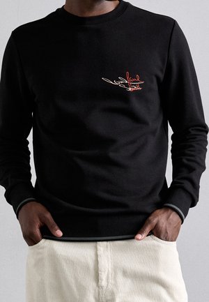 Mężczyzna w czarnej koszuli z długim rękawem z czerwono-białym haftowanym logo "Paul Smith" na piersi i jasnobeżowych spodniach sztruksowych, ręce w kieszeniach.