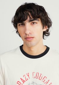 Tricou alb cu mânecă scurtă și guler negru, cu imprimeu roșu pe care scrie "CRAZY COUGARS". Modelul are păr ondulat de culoare închisă și o expresie neutră.