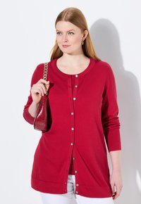 Roter Cardigan mit rundem Ausschnitt, langen Ärmeln und Knopfleiste. Getragen mit weißen Hosen und einer kleinen roten Handtasche.