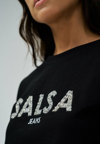 Sudadera negra con la palabra "SALSA" en lentejuelas plateadas y "JEANS" bordado debajo. Diseño de cuello redondo con una textura suave.