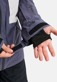 Paarse waterdichte jas van Gore-Tex. Voorzien van verstelbare klittenbandmanchetten en een zwarte binnenvoering met duimopeningen. Toont een soepele textuur.