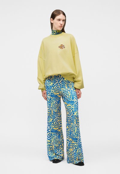 Femme portant un sweat-shirt jaune oversize et un pantalon large à motifs bleu, jaune et blanc, debout devant un fond uni.