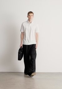 Λευκό κοντομάνικο πόλο πουκάμισο, μαύρο παντελόνι με φαρδιά μπατζάκια, κρατώντας μια μαύρη τσάντα, στέκεται σε λείο γκρι πάτωμα μπροστά σε απλό λευκό φόντο.