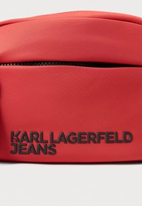 Czerwona nylonowa saszetka z czarnym suwakiem, z wytłoczonym napisem „KARL LAGERFELD JEANS” na przedzie jako znak firmowy. Gładka tekstura, minimalistyczny design.