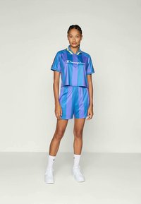 Žena oblečená v modro-fialovom pruhovanom športovom súprave Champion, skladajúcej sa z kraťasov a topu, s bielymi ponožkami a bielymi teniskami, stojí na jednoduchej pozadí.