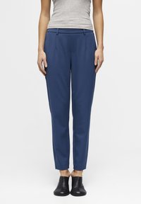 Een persoon staat gekleed in een grijze tanktop en blauwe broek, gecombineerd met zwarte instapschoenen; schone, minimalistische achtergrond.
