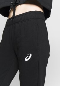 Zwarte trainingsbroek met een elastische tailleband, voorzien van een wit logo aan de linkerkant. Het materiaal lijkt zacht en glad met een comfortabele pasvorm.