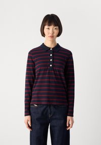 Woman wearing a navy and burgundy striped long-sleeve polo shirt with gold buttons and dark blue jeans, standing against a plain background.