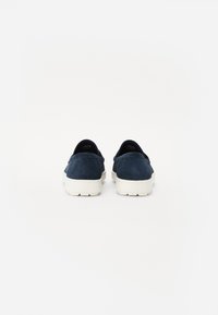 Chaussures slip-on en daim bleu marine avec une semelle en caoutchouc blanc, présentant des talons arrondis et des accents minimaux, une surface texturée et aucune lacet visible.