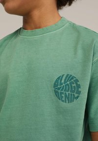 Žalias medvilnės marškinėliai su apvaliu jūros mėlynumo grafiniu dizainu, kuriame yra tekstas "ALL RIDE DENIM." Klasikinis apvalus iškirpimas ir trumpomis rankovėmis.