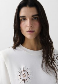 Felpa bianca con un intricato design ricamato a forma di sole sulla spalla e dettagli ritagliati delicati. Texture liscia e vestibilità casual.