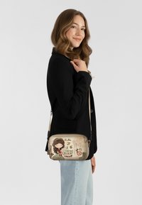 Umhängetasche mit einer beigen Stoffoberfläche, die ein Cartoon-Design zeigt, hellen Beschlägen und einem verstellbaren Riemen, ergänzt durch eine schlanke rechteckige Form.