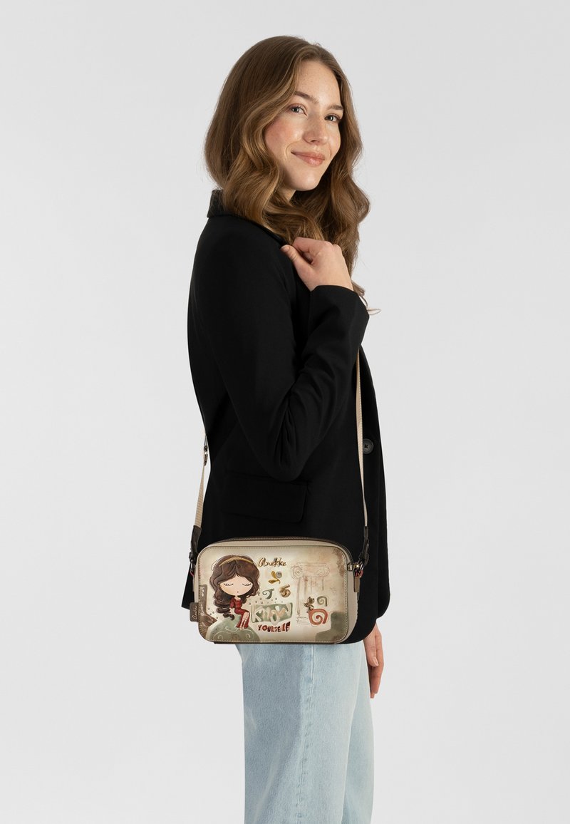 Umhängetasche mit einer beigen Stoffoberfläche, die ein Cartoon-Design zeigt, hellen Beschlägen und einem verstellbaren Riemen, ergänzt durch eine schlanke rechteckige Form.