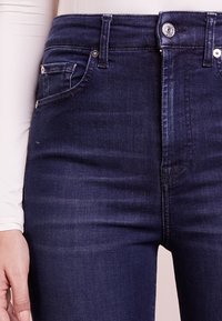 Los jeans de mezclilla azul oscuro cuentan con un diseño ajustado, estilo de cinco bolsillos y un cierre de botón. La tela tiene una textura suave con un ligero desgaste.