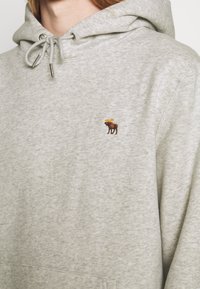 Grijze fleece hoodie met een kangoeroezak, trekkoordkap en een kleine bruine eland borduursel op de linkerkant van de borst.