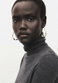 Top de cuello alto gris con textura suave, combinado con grandes pendientes de aro dorados, con un retrato en primer plano sobre un fondo neutro.