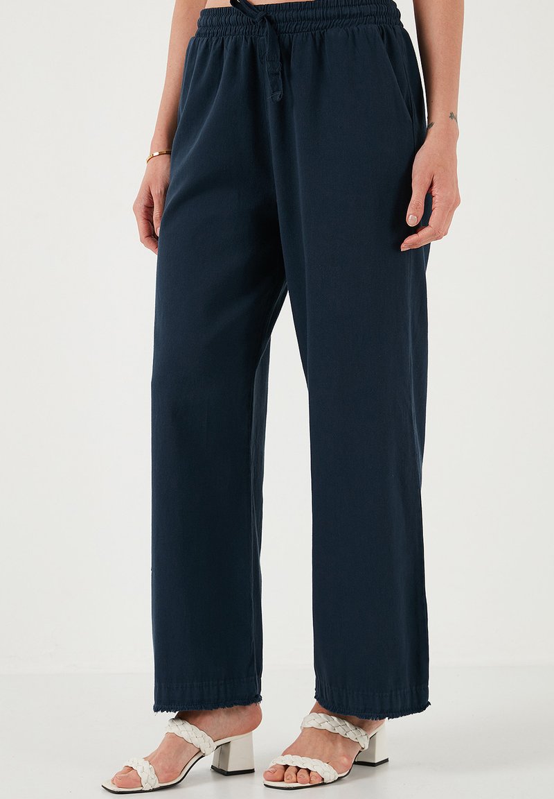 Donkerblauwe wijduitlopende broek met een elastische tailleband en koord. Zachte stof met een ontspannen pasvorm en rafelige zoom. Gepaard met witte haksandalen.