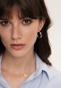 Złote kolczyki w kształcie kół, delikatny złoty naszyjnik z okrągłym i sercowym wisiorkiem, noszone na jasnoniebieskiej koszuli z kołnierzykiem. Gładkie metaliczne wykończenie.