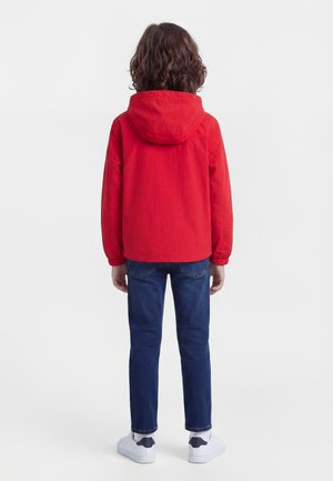 Barn med krøllete hår iført en rød hettejakke, blå jeans og hvite joggesko, stående med ryggen mot på hvit bakgrunn.