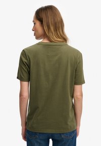 Camiseta de manga corta color verde oliva con cuello redondo, textura suave de algodón y un detalle de parche cuadrado en la parte superior de la espalda.
