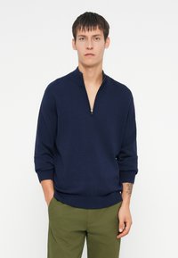 Granatowy sweter z zamkiem i długimi rękawami, z prążkowanymi mankietami i dołem, wykonany z miękkiego materiału. Nosi się go z oliwkowymi spodniami.