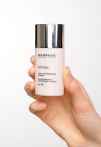 Bouteille rose légère avec un bouchon argenté, étiquetée "DARPHIN INTRAL", contenant un écran environnemental SPF 50 ; tenue par une main.