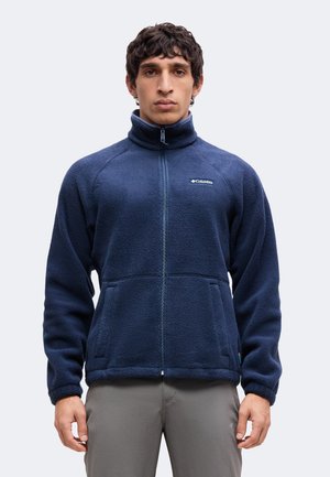 Homme portant une veste polaire bleu marine avec une fermeture éclair à l’avant et deux poches, debout devant un fond clair uni.
