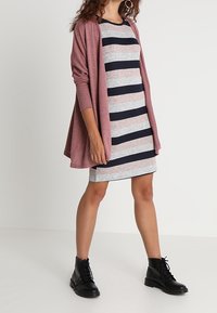 Cardigan aperto color rosa sopra un vestito fitted a righe blu navy e grigio. L'outfit è abbinato a stivaletti neri con lacci alla caviglia.