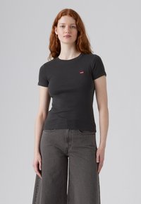 Zwarte t-shirt met korte mouwen van katoen, met een klein rood Levi's-logo op de borst. Draagt bij losse grijze wijde jeans.