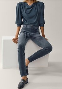 Kvinna som sitter på vit block klädd i en mörkblå draperad blus, högmidjade sammetbyxor och svarta loafers med metalldetaljer.