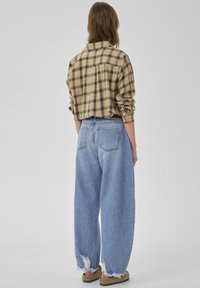 Flannelskjorte med tern i tan og mørkebrun, kombineret med lyseblå vidde bukser med frynser. Modellen står med ryggen til.