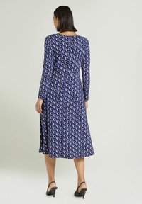 Sukienka z długim rękawem, niebieska z geometrycznym białym wzorem, do kolan, o gładkiej fakturze i dopasowanej talii. Noszone czarne sandały na obcasie.