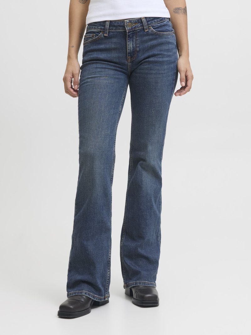 Pessoa vestindo jeans bootcut azuis e sapatos de couro pretos, de pé contra um fundo branco liso.