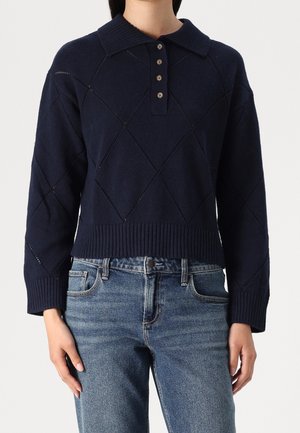 Granatowy sweter dziergany z prążkowanym kołnierzem, z plisą na guziki i wzorem rombów. W połączeniu z wystrzępionymi niebieskimi dżinsami.