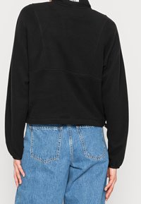 Musta lyhyt huppari, jossa on korkea kaulus ja saumadetalji selän yläosassa, yhdistetty sinisiin korkeavyötäröisiin farkkuihin.