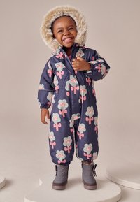 Combineu de zăpadă izolat, bleumarin, cu un model floral în culori pastel, având o glugă captusită cu blană artificială și manșete elastice, purtat cu bocanci gri.