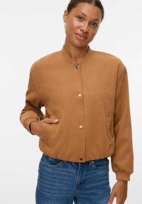 Femme aux cheveux courts et bouclés portant une veste beige à boutons et un jean bleu, se tenant avec une main dans la poche sur fond blanc.