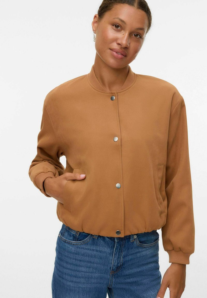 Femme aux cheveux courts et bouclés portant une veste beige à boutons et un jean bleu, se tenant avec une main dans la poche sur fond blanc.