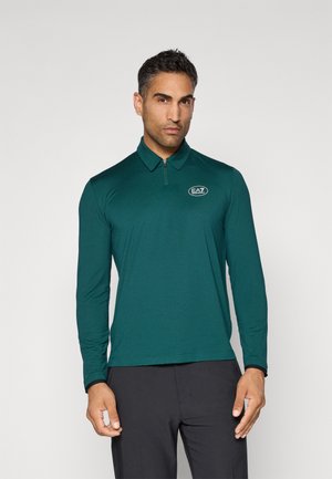 Man wearing dark green long-sleeve polo shirt with zipper and white EA7 logo on chest, paired with black pants, standing against plain background.