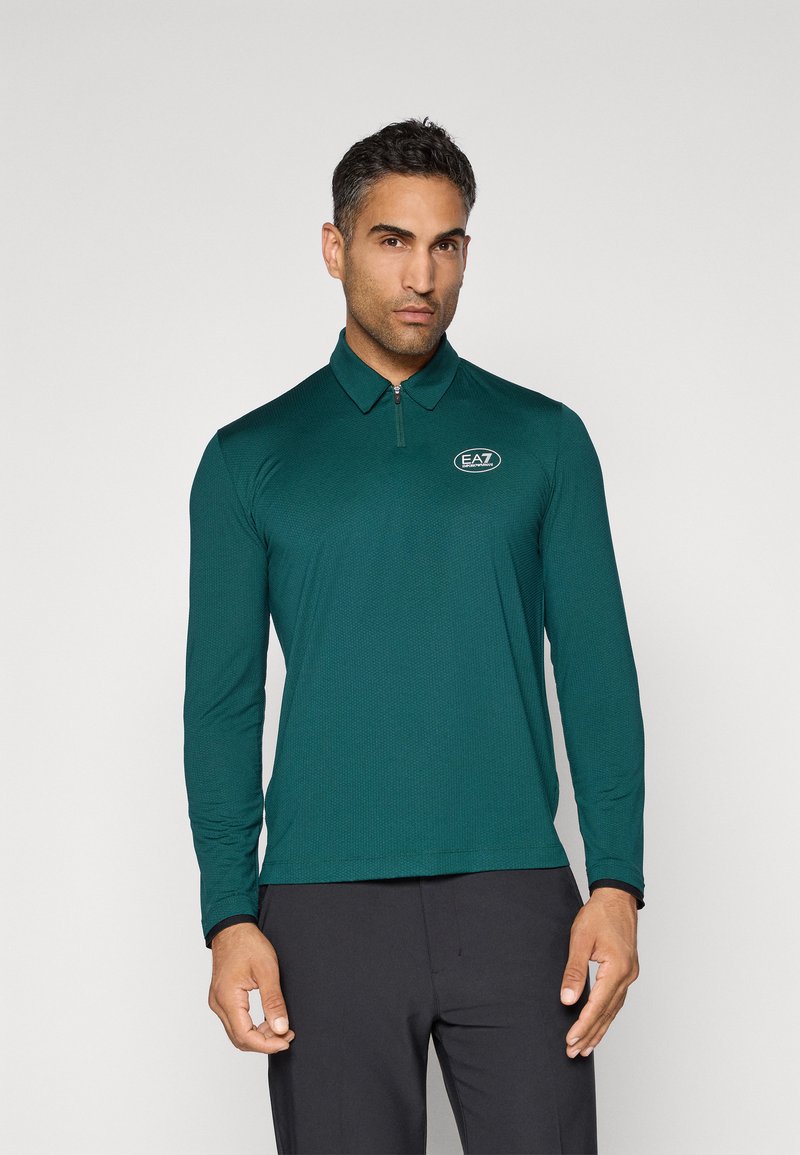 Mann som har på seg en mørkegrønn langermet polo med glidelås og hvit EA7-logo på brystet, iført svarte bukser, står foran en ensfarget bakgrunn.