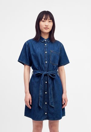 Frau trägt ein kurzärmliges blaues Jeanskleid mit Knöpfen vorne, Brusttasche und gebundenem Taillengürtel, steht vor einem schlichten weißen Hintergrund.