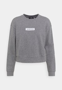 Sweatshirt gris court en tissu doux. Présente des poignets et un ourlet côtelés, avec une étiquette rectangulaire blanche portant la mention "CALVIN KLEIN" sur le devant.
