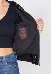 Persoon draagt een zwart crop top en blauwe jeans, opent een zwarte bomberjas met pailletten en toont het binnenlabel met oranje tekst.