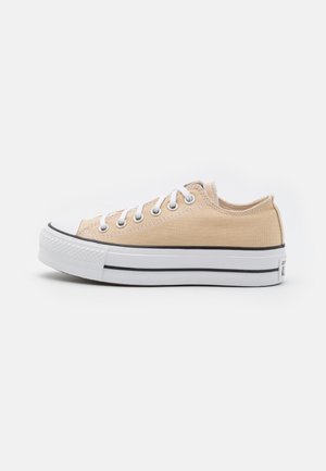 Beige lavt canvas sneakers med hvid gummisål, hvide snørebånd og sort stribe på sålen mod hvid baggrund.