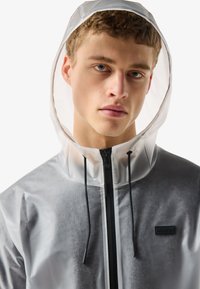 Young man with short curly hair wearing translucent gray hooded rain jacket with black zipper and drawstrings, neutral background.