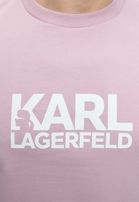 Ροζ μπλουζάκι με μεγάλο λευκό κείμενο "KARL LAGERFELD" και μικρή σιλουέτα ατόμου με γυαλιά ηλίου και αλογοουρά μέσα στο γράμμα "K".
