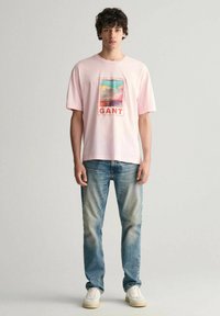 Lichtroze T-shirt met korte mouwen, voorzien van een kleurrijke grafische print met het woord "GANT", gecombineerd met vervaagde blauwe jeans en witte sneakers.