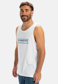 Biely bavlnený top na ramienka s modro-čiernym logom v obdĺžnikovom dizajne. Materiál je hladký a strih je voľný. Pod ním sú oblečené džínsy.